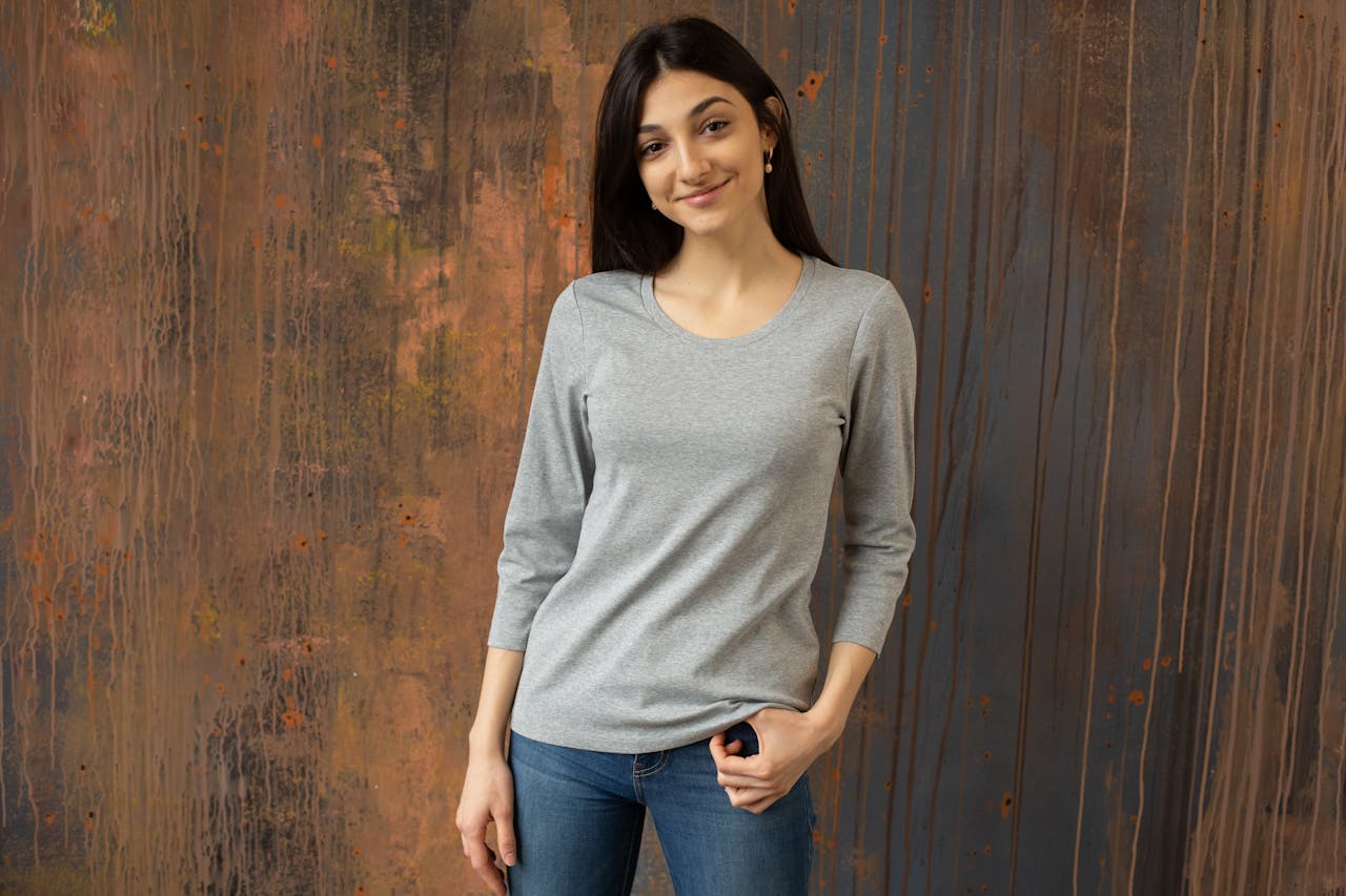 Young glad ethnic female in jeans and gray top standing against shabby wooden wall and looking at camera contentedly