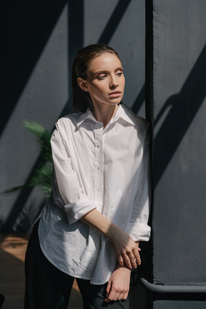 A woman in a white shirt leans thoughtfully against a shadowed wall indoors.